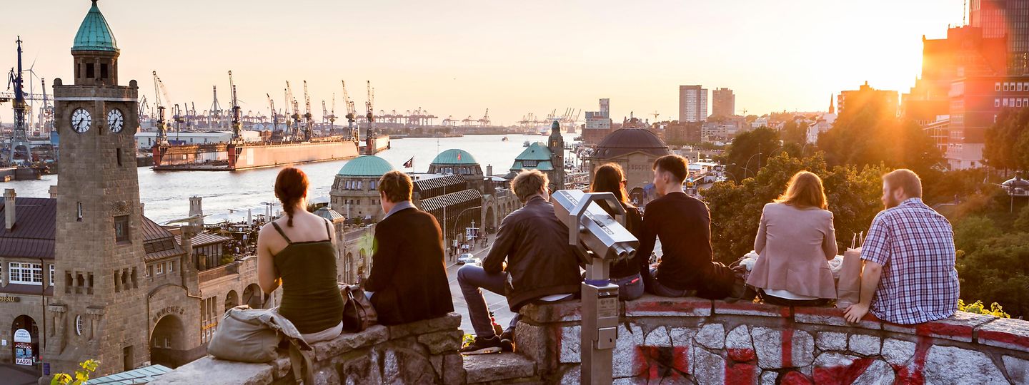Junge Menschen sitzen bei Abendstimmung auf einer Mauer und blicken Richtung Landungsbrücken