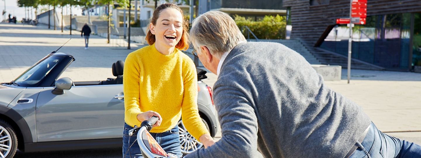Eine Frau hilft einem Mann auf ein StadtRAD, während ihr gerade geliehenes Cabrio von SIXT Share im Hintergrund geparkt ist